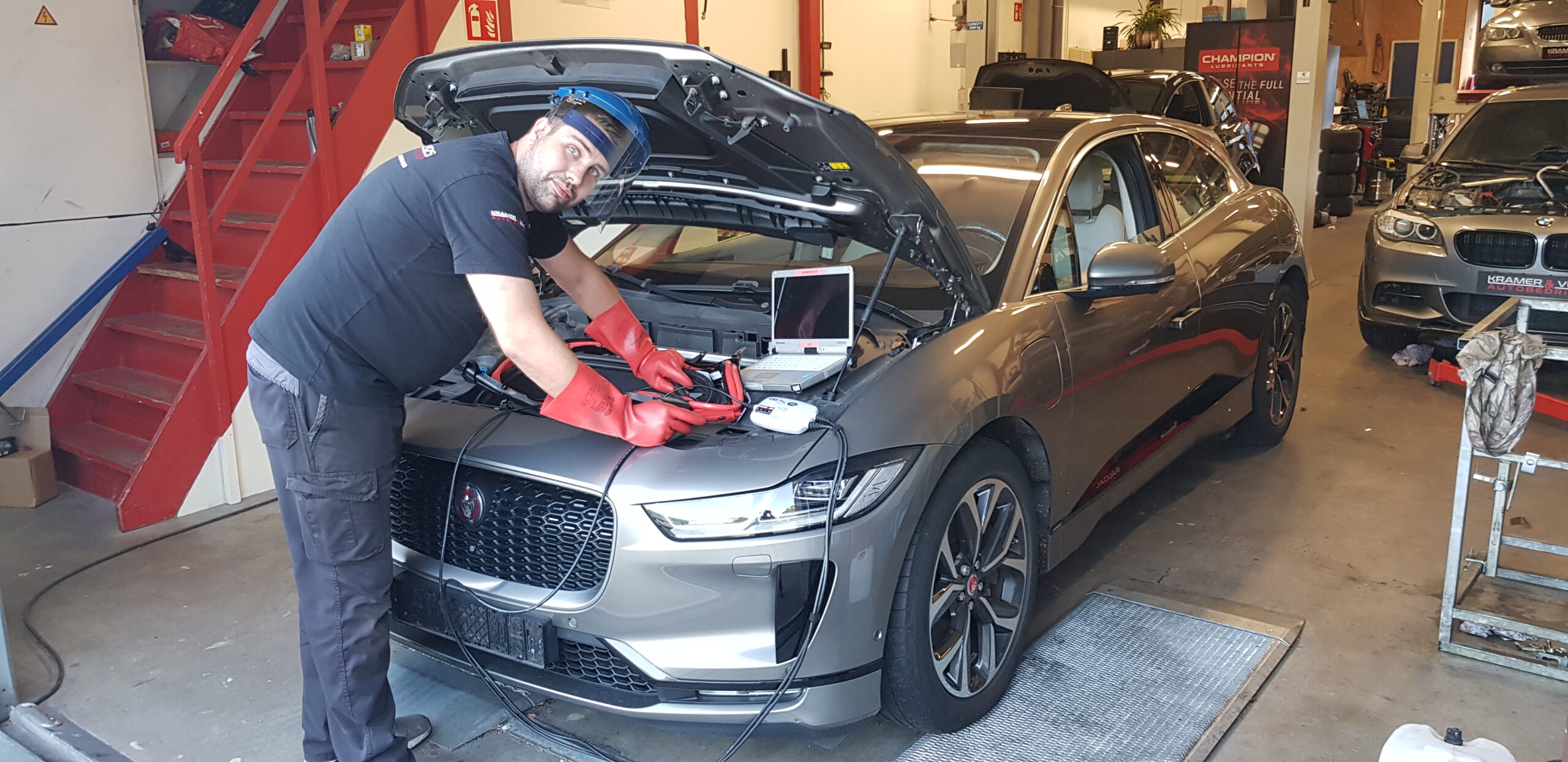 Michel voert met beschermende kleding en een gelaatsscherm een softwarematige diagnose uit aan de motorruimte van een grijze Jaguar I-Pace met behulp van een laptop.